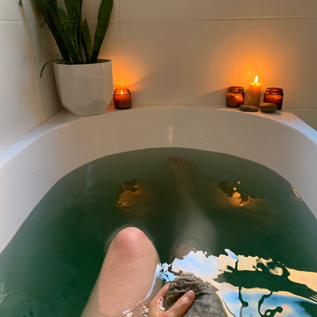 Person soaking in a magnesium herbal bath thats green with candles and a plant